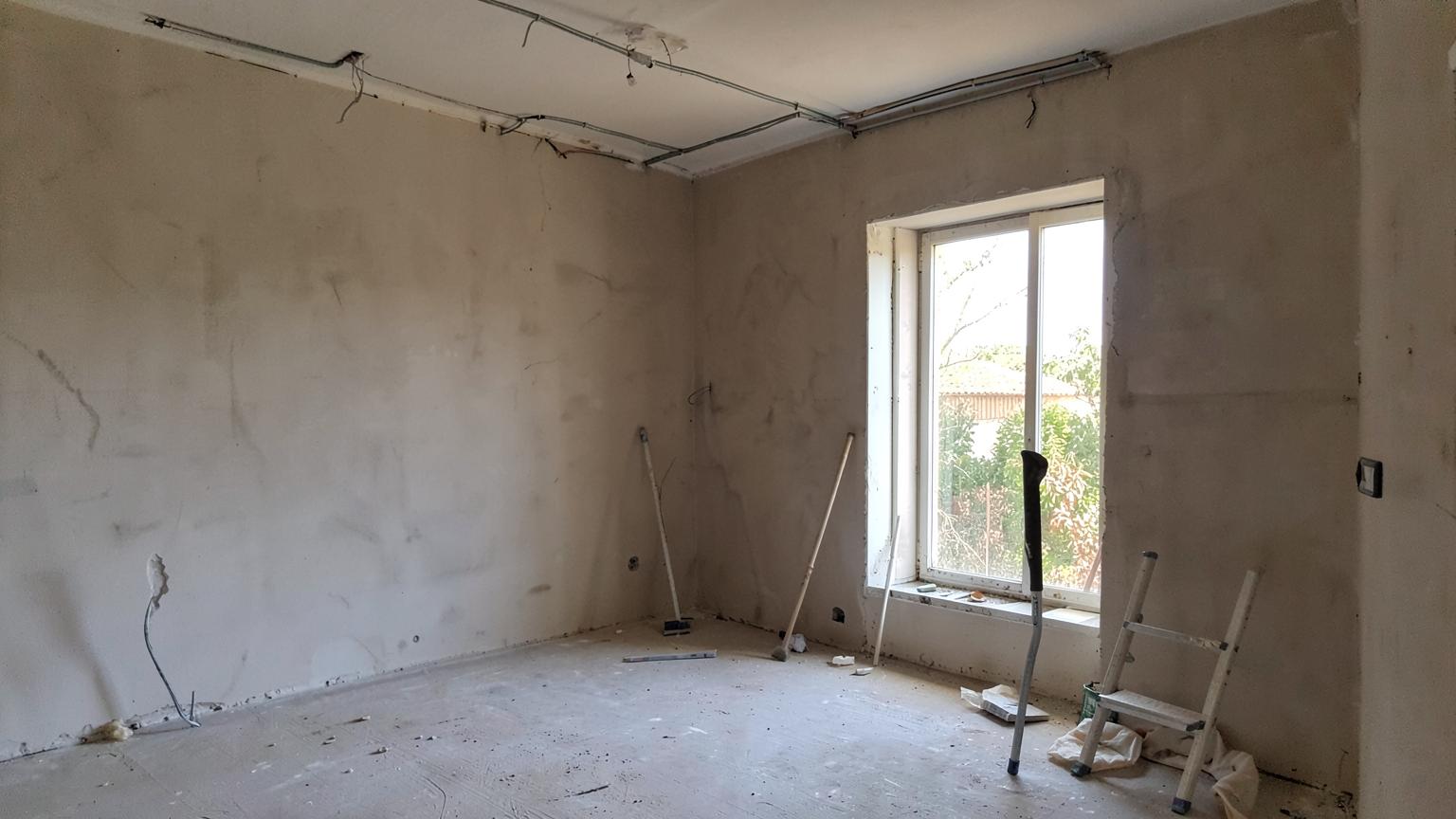 Empty villa room mid-renovation with bare plaster walls, exposed conduit, ladder and dust sheets by a large window